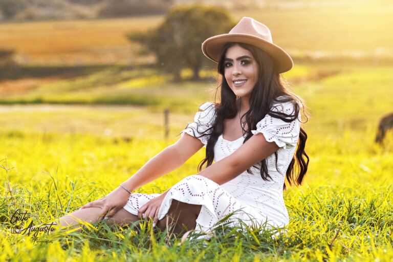 Retrato feminino em campo ao pôr do sol, com vestido branco e chapéu, destacando a beleza natural e a luz suave do final do dia.