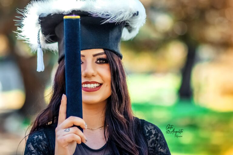 Mulher sorrindo em traje de formatura, segurando diploma em um parque ao ar livre.