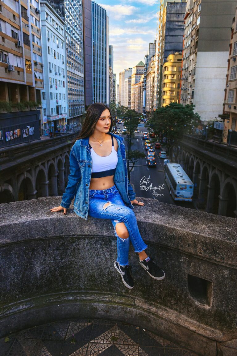 Jovem mulher sentada em uma ponte no centro de Porto Alegre, usando jaqueta jeans e calça rasgada, com expressão contemplativa. Ao fundo, prédios e o movimento urbano da cidade.
