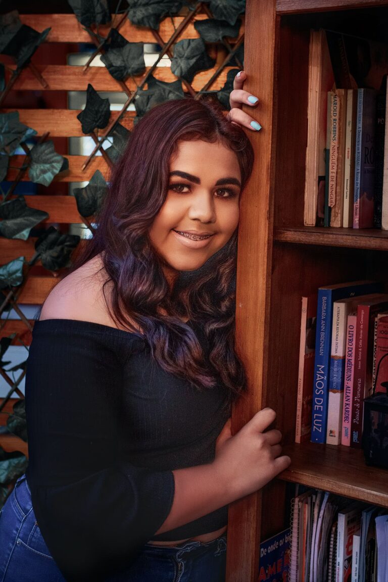 Ensaio fotográfico de 15 anos de Alana em Guaíba, junto à estante de livros, capturando seu sorriso e expressão autêntica