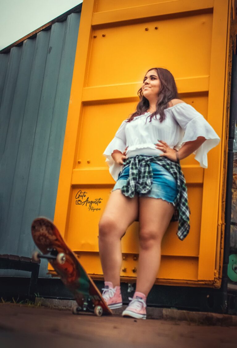 Alana em ensaio de 15 anos com tema urbano, posando com skate em frente a estrutura amarela em Guaíba