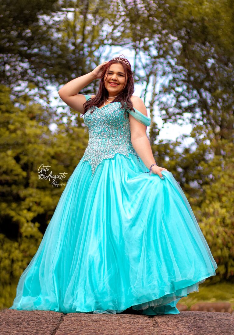 Alana sorrindo em seu ensaio de 15 anos, usando um vestido azul brilhante e posando ao ar livre em um cenário natural. Fotografia por Guto Augusto.