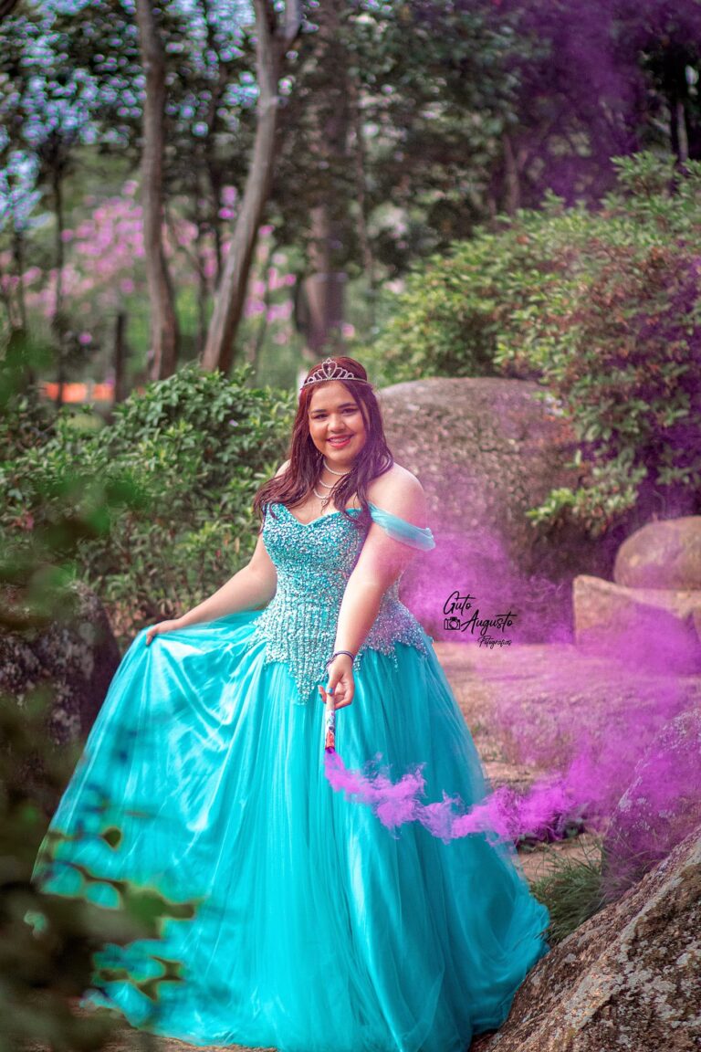 Alana em seu ensaio de 15 anos, sorrindo e segurando uma fumaça colorida em meio à natureza, usando um vestido azul brilhante. Fotografia por Guto Augusto.