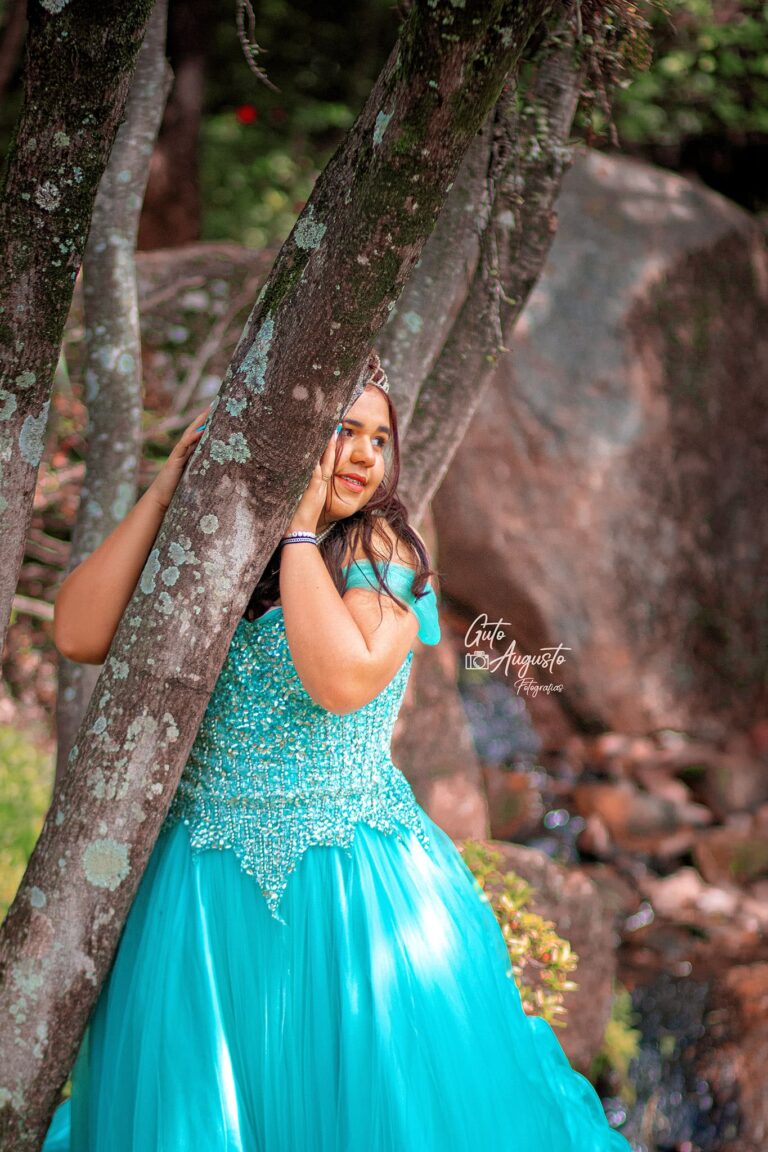 Alana sorrindo em seu ensaio de 15 anos, usando um vestido azul brilhante e posando ao ar livre em um cenário natural. Fotografia por Guto Augusto.