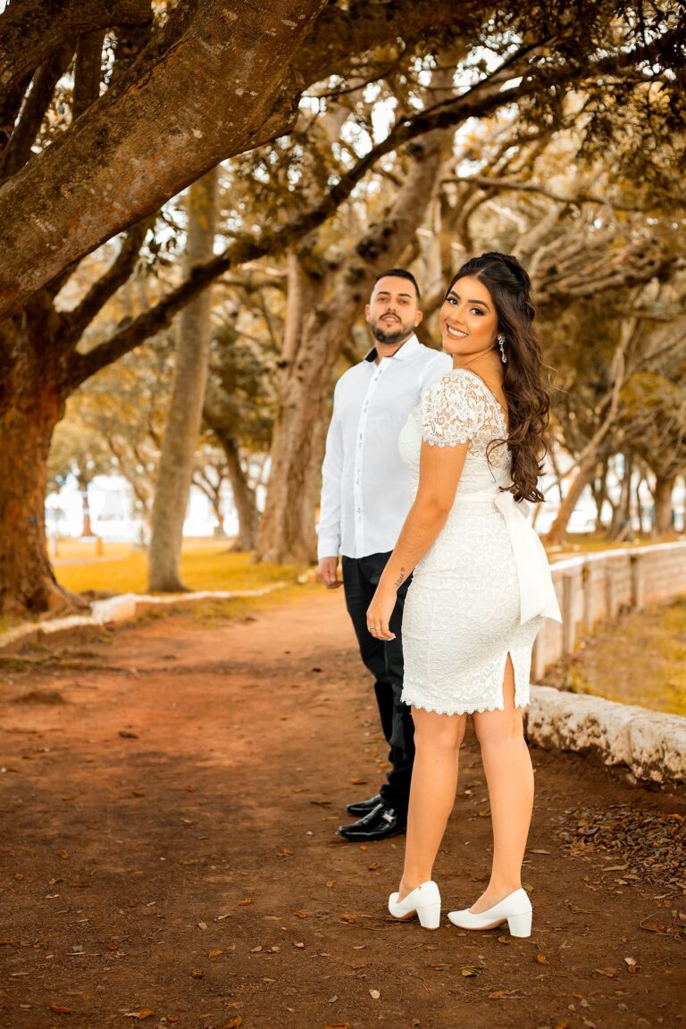 Casal sorridente em parque arborizado, mulher em vestido branco de renda, homem ao fundo em camisa azul clara, cenário romântico.